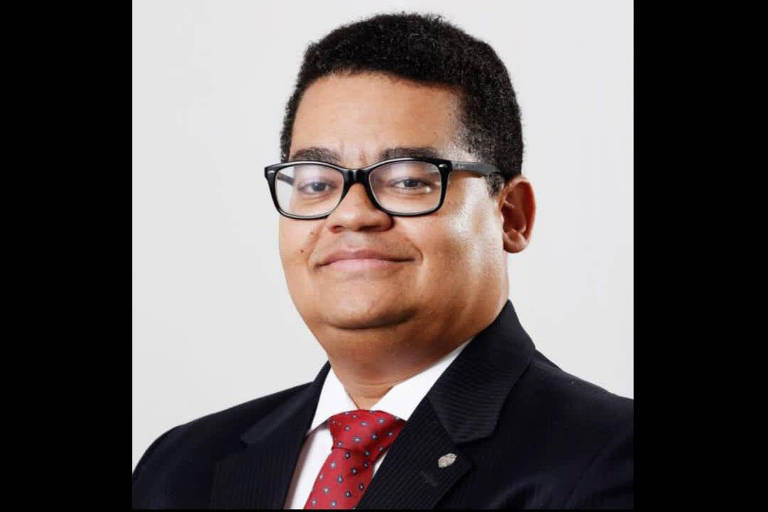 Rodrigo Monteiro é um homem preto, usando paletó preto, camisa branca, gravata vermelha e óculos