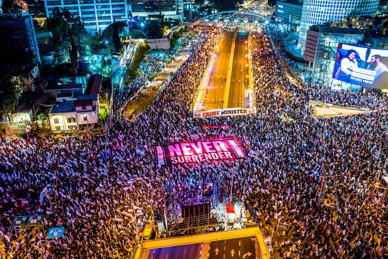 a fotografia aérea mostra uma multidão que toma as ruas de tel aviv. no centro da imagem, iluminado por uma luz roxa, está escrita a expressão "never surrender" (nunca se renda).