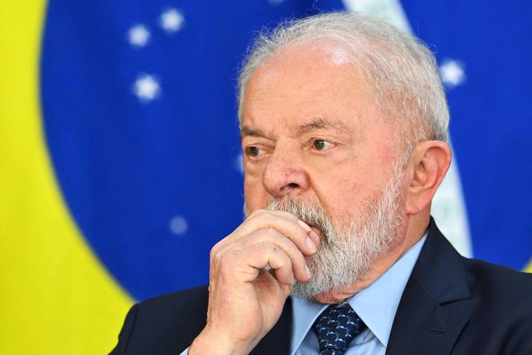 Presidente Lula, em close, com a  mão na barba; ao fundo, a bandeira do Brasil