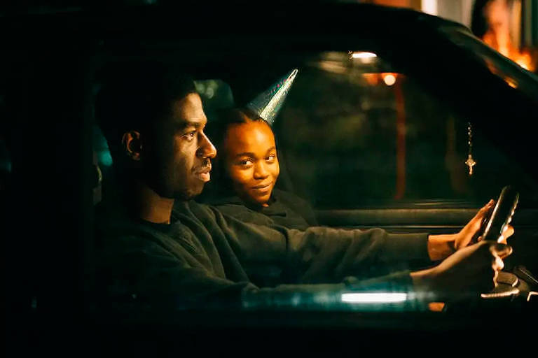 Em cena do thriller "Excluídos", um homem dirige um carro ao lado de uma mulher que usa um chapéu de aniversário e sorri