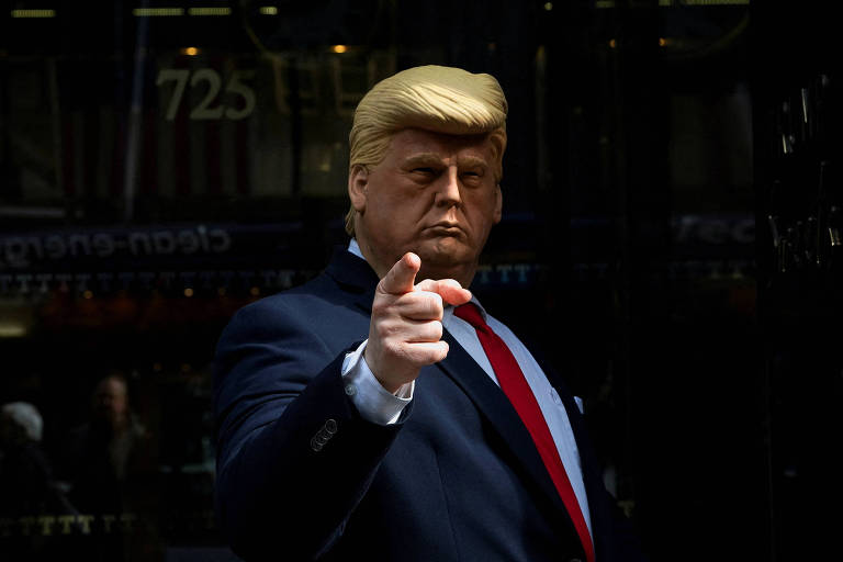 pessoa com uma máscara facial de donald trump aponta o dedo para a câmera em frente a um edifício. ele usa terno azul e gravata vermelha. a feição de trump na máscara é séria