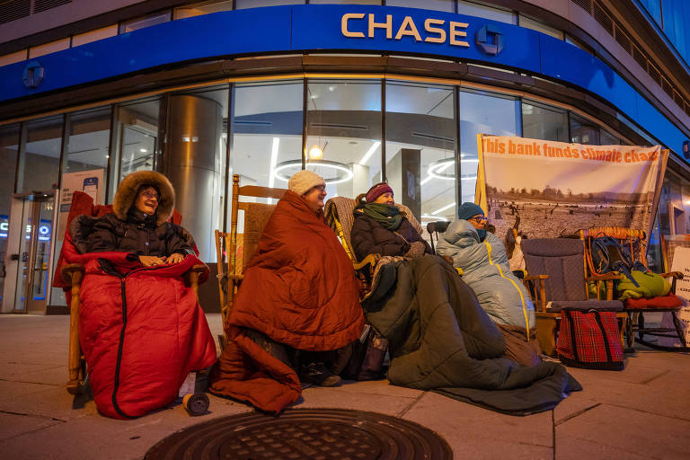 Idosos sentados com cobertores, edredons e cartazes na calçada em frente à fachada do banco