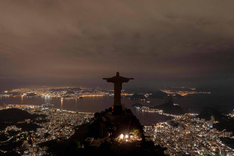 Cristo no escuro, no Rio, durante a noite, com luzes em outros pontos da cidade