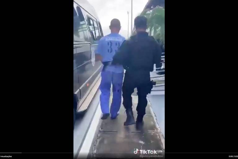 Policial leva detento pelo braço esquerdo. Os dois aparecem de costas, caminhando, e há o que parece ser uma van ou ônibus à esquerda. Estão em local a céu aberto