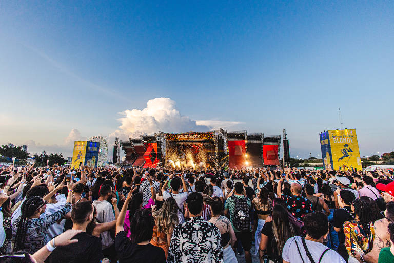 A imagem mostra uma grande multidão em um show ao ar livre, com pessoas levantando as mãos em direção ao palco. O céu está claro, com algumas nuvens, e o palco é visível ao fundo, decorado com luzes e banners. À esquerda e à direita, há placas coloridas 