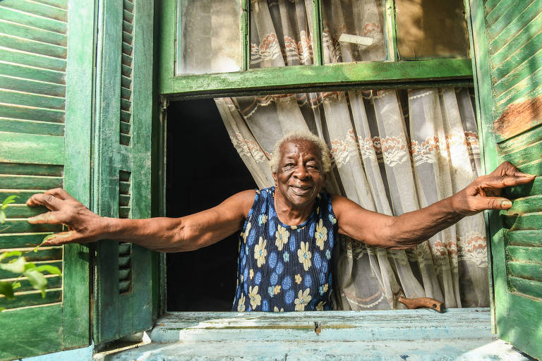 Mulher negra e idosa posa para foto; ela aparece abrindo a janela de casa e sorrindo
