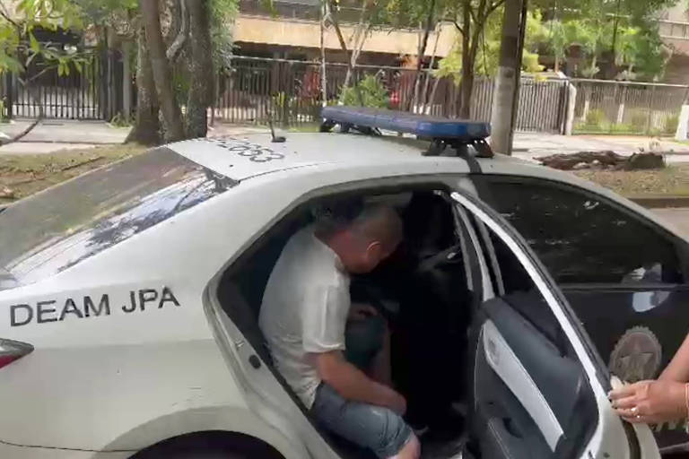 Um homem está saindo de um carro de polícia. Ele usa camiseta branca e não é possível ver seu rosto