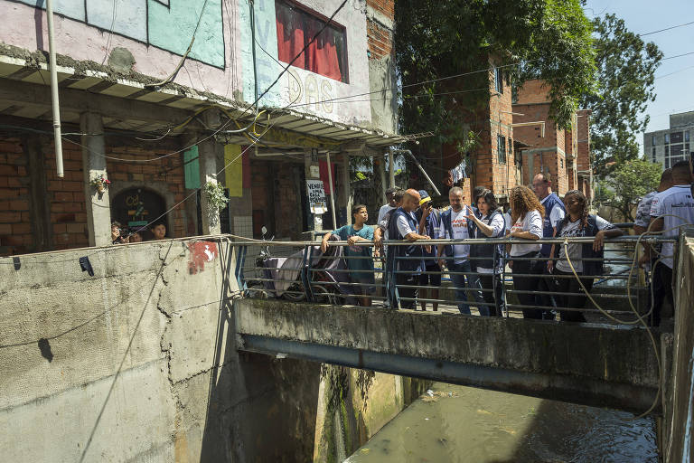 Dezenas de pessoas se aglomeram em uma pequena monte dentro de uma favela. Por baixo da estrutura passa uma água suja e turva