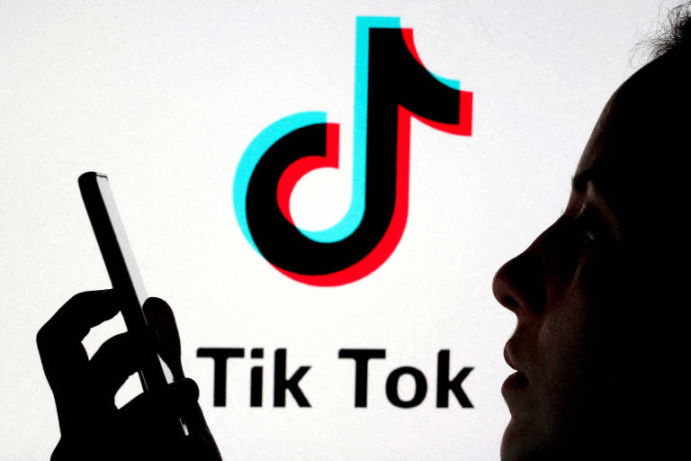 Foto mostra logo do TikTok, uma colcheia preta contornada por azul e vermelho, posicionada entre o rosto e o celular da uma silhueta de uma pessoa