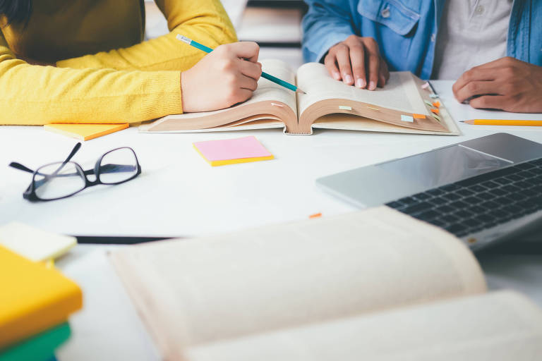 A imagem mostra duas pessoas estudando juntas em uma mesa. Uma delas está escrevendo em um caderno com um lápis verde, enquanto a outra está lendo um livro. Há um laptop aberto, óculos, post-its coloridos e vários livros espalhados pela mesa.