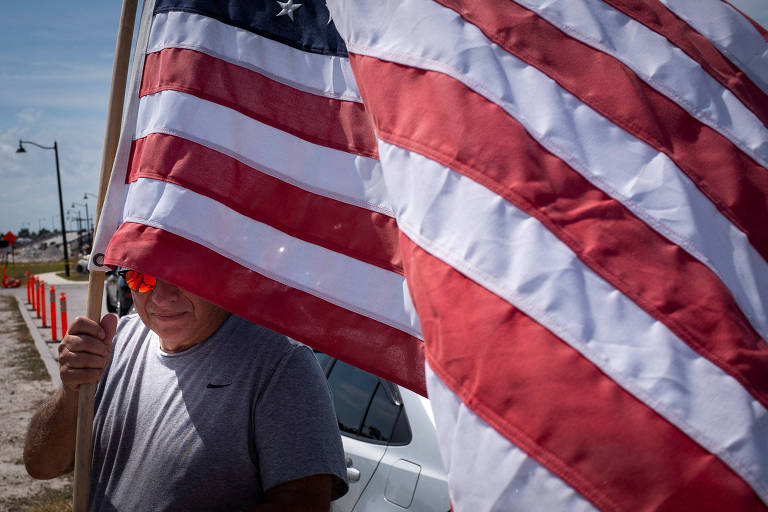 a foto mostra um homem branco de meia idade, que usa óculos alaranjados, parcialmente coberto pela grande bandeira dos estados unidos que segura em mãos
