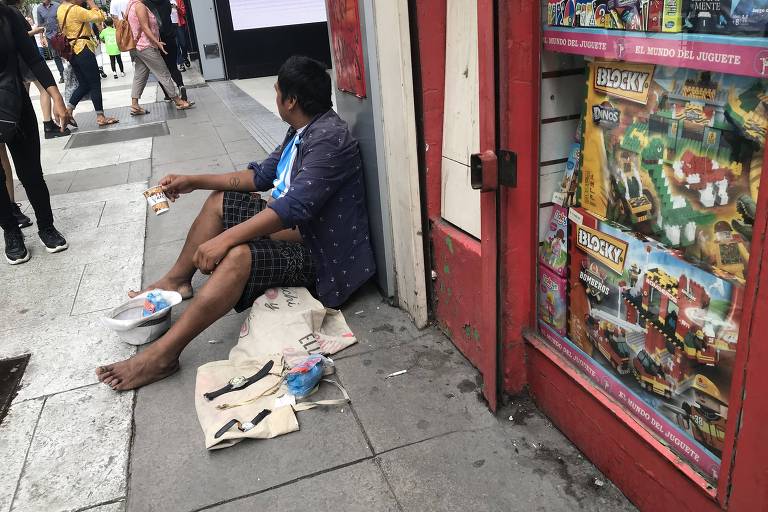 Morador de rua está decalço e sentado em uma calçada. Ele se apoia na marquize de uma loja.