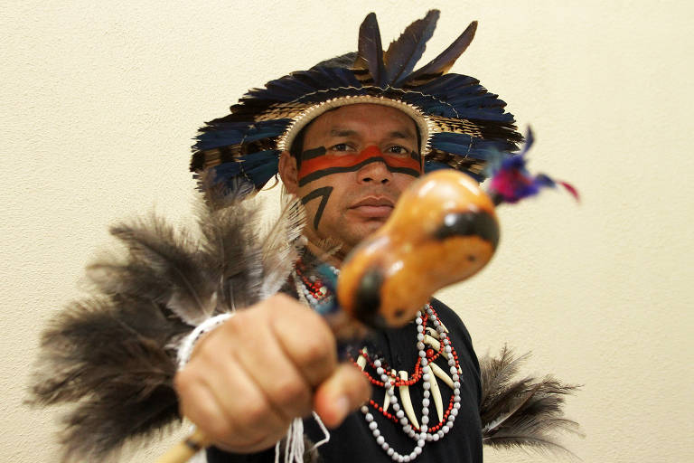 Homem indígena veste camiseta preta e artefatos indígenas, como colares, cocar e um instrumento na mão direita, que ele coloca à frente
