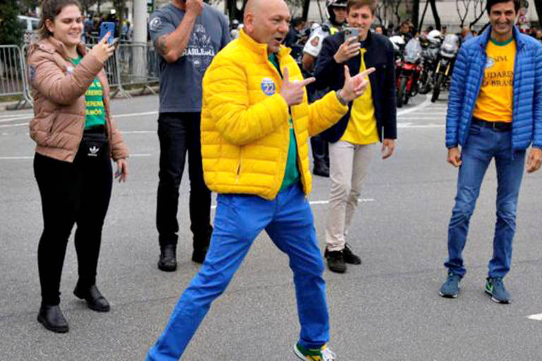 Luciano Hang se veste de jaqueta puffer amarela, calças jens e camiseta verde. Por detrás dele, estão fãs. Hang é influenciador digital, além de dono da Havan.