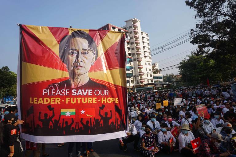 a foto mostra uma manifestação que reúne milhares de pessoas nas ruas de Rangoon. um grande cartaz está erguido com o rosto de Aung San Suu Kyi, uma mulher idosa asiática. lê-se, no cartaz, em inglês: por favor, salvem nossa líder, nosso futuro e nossa esperança