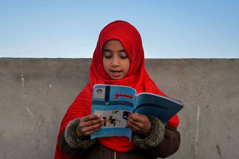 Fotografia colorida em plano médio de criança muçulmana; ela usa um véu vermelho em volta da cabeça e lê um livro