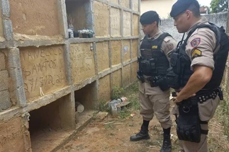 Imagem colorida mostra dois policiais militares em frente ao túmulo aberto do cemitério de Visconde do Rio Branco, onde uma mulher foi resgatada viva