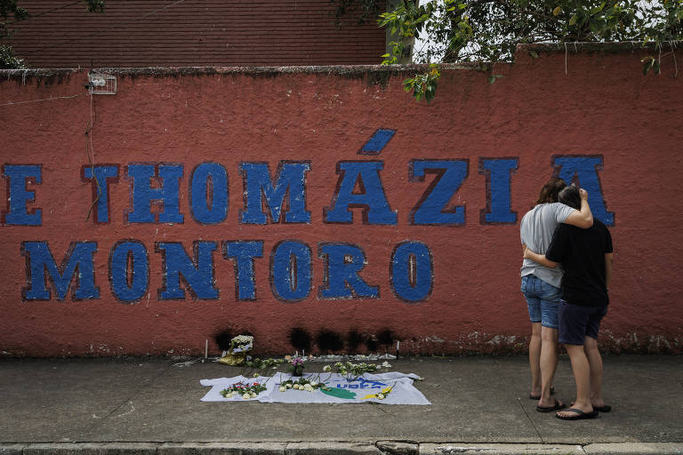 Duas mulheres abraçadas em frente à escola e próximas a ponto na calçada onde foram deixadas velas e flores 