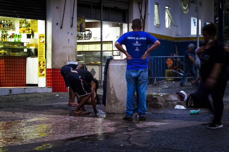 foto mostra pessoas transitando e aguardando para usar fonte de água, enquanto dois homens estão agachados lavando roupa no equipamento 