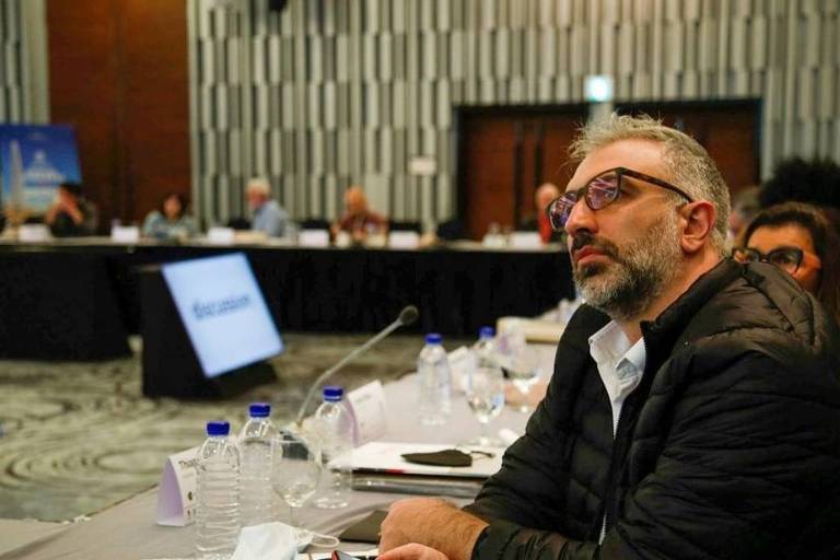 Homem grisalho de barba e óculo sentado em uma mesa de conferência, aparece de perfil