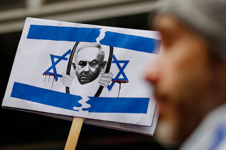 manifestante exibe o cartaz de uma bandeira de israel modificada. nela, aparece a imagem do primeiro-ministro Netanyahu atrás das grades, enquanto escorre sangue da estrela de davi