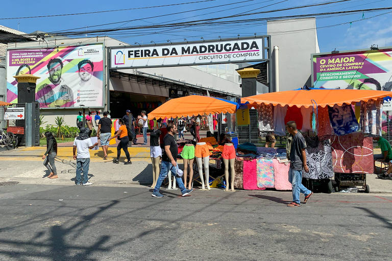 Fachada do shopping Feira da Madrugada, no Brás, no centro de São Paulo, com barracas de toldo laranja de ambulantes vendendo roupas coloridas na entrada