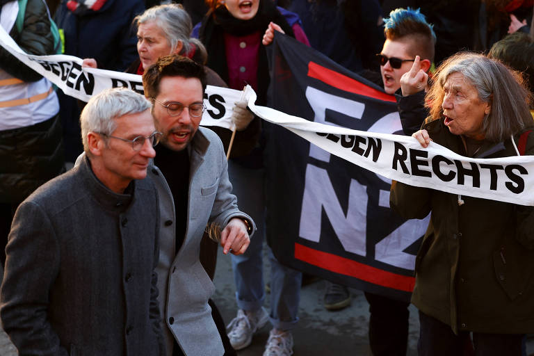 enquanto dois homens passam na rua, manifestantes carregando cartazes antinazistas gritam e fazem gestos obscenos em sua direção
