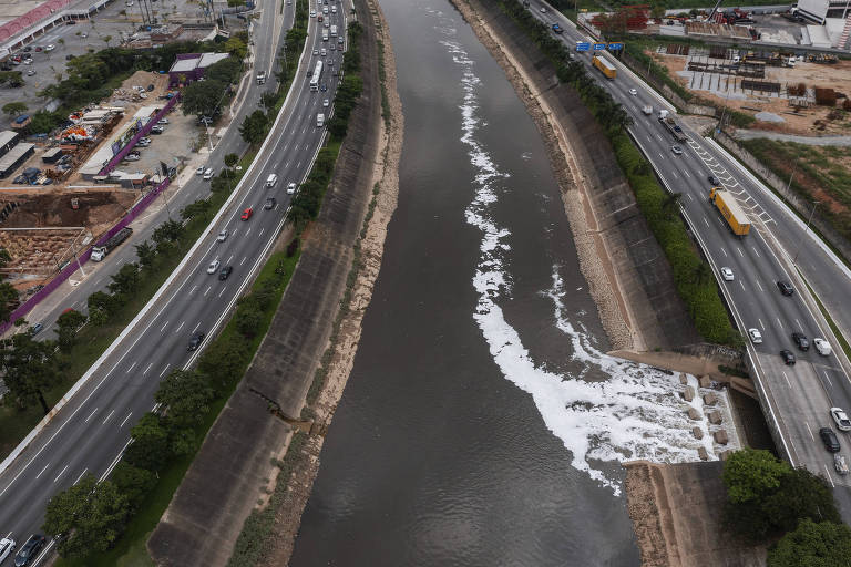 Vista aérea mostra águas poluídas de afluente sendo jogadas no rio Tietê, formando uma mancha de espuma branca sobre as águas escuras do rio canalizado e cercado por avenidas marginais