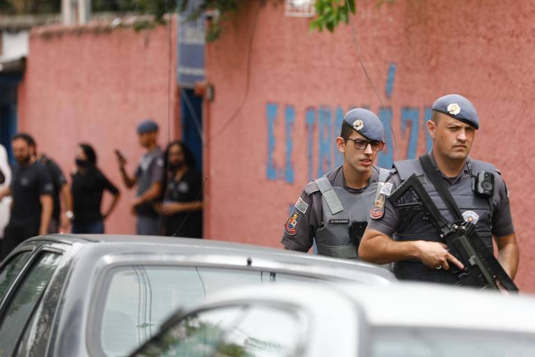 Policiais militares, um deles portando fuzil, em primeiro plano em frente a muro laranja de escola estadual