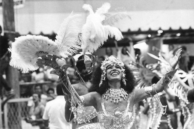 Foto em preto e branco mostra mulher em desfile de carnaval  com as mãos para cima, sorriso aberto e coroa de conchas na cabeçala