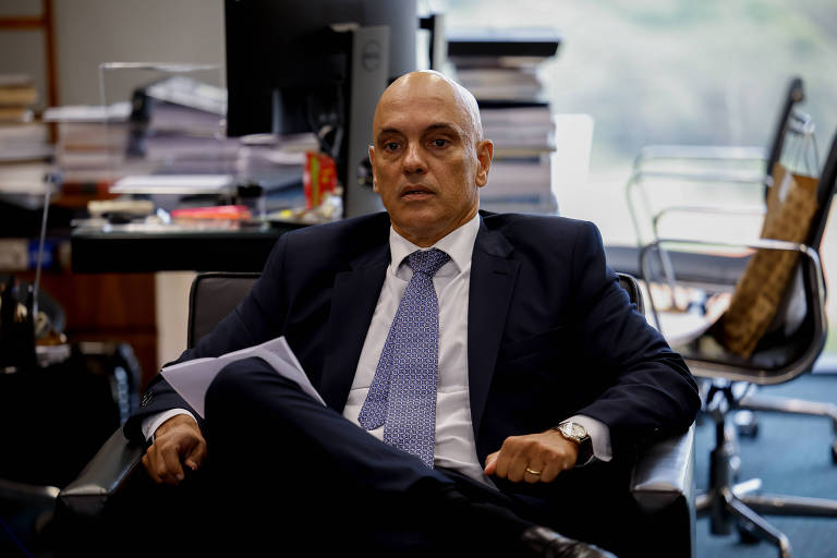 Alexandre de Moraes, homem branco, está sentado em cadeira em sala com livros e documentos. Ele usa camisa branca, gravata azul e terno escuro