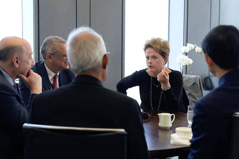 Dilma, uma mulher idosa de cabelos claros e curtos, aparece na ponta de uma mesa e é fotografada falando enquanto é observada por outros quatro homens que estão em reunião. todos usam roupas formais