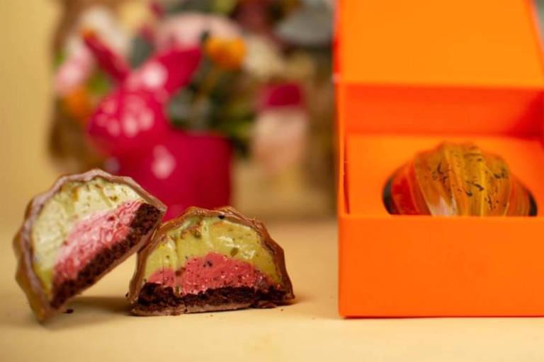 A foto mostra uma caixa com um chocolate em formato de cacau. Ao lado, o chocolate aparece cortado revelando o recheio em duas cores: verde (pistache) e rosa (frutas vermelhas)