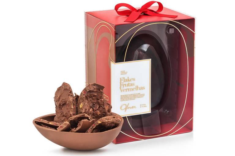 A foto traz uma casca de chocolate com pedaços de casa recheada com flocos de milho crocantes e frutas vermelhas secas. Ao fundo, uma caixa vermelha com laço