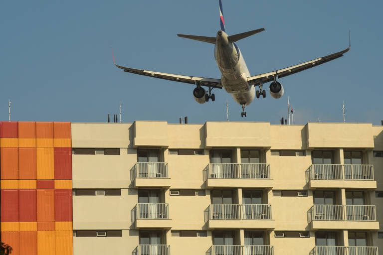 Avião passa perto do topo de prédio que possui varandas e uma das laterais decorada com painéis nas cores amarelho, vermelho e laranja