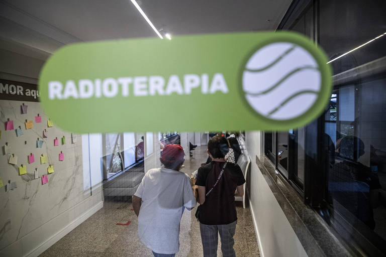 Duas pacientes, de costas para a foto, caminham no corredor do Hospital Haroldo Juaçaba. Numa placa em primeiro plano, lê-se radioterapia
