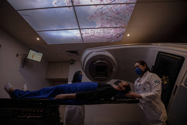 Paciente deitado na máquina de radioterapia, com profissional de saúde atrás dele posicionando-o para início da sessão; no teto, há imagem de flores  