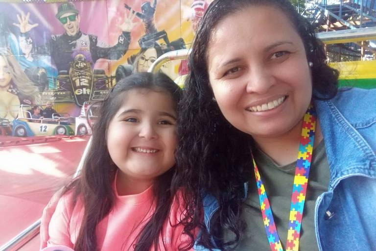 Foto colorida de Marina e Erika sorrindo. A menina tem cinco anos, está à esquerda. É branca, tem cabelo liso e comprido. Erika, à direita, é morena, tem cabelo enrolado e usa um cordão colorido com quebra-cabeças nas cores amarela, vermelha, azul e verde. 