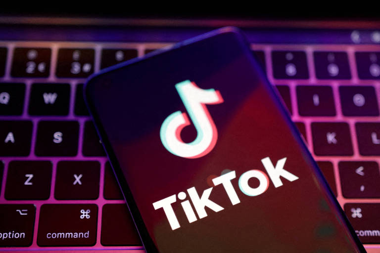 Celular preto com símbolo do do TikTok escrito em branco na tela; ao fundo, há um teclado preto