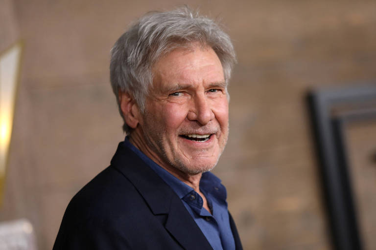 O ator Harrison Ford, um homem grisalho, sorri para a lente da câmera e usa terno preto e camisa azul