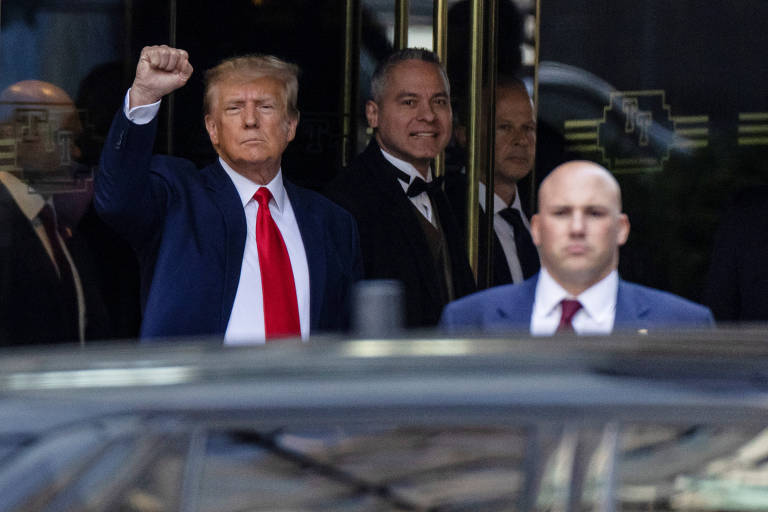 Com camisa branca, gravata vermelha e terno azul escuro, Trump ergue o punho aberto atrás de um carro, em primeiro plano na foto