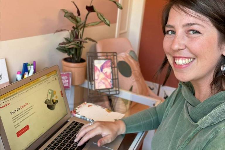 mulher branca com blusa verdade sorri em frente a computador na sua mesa, que tem flores e um quadro