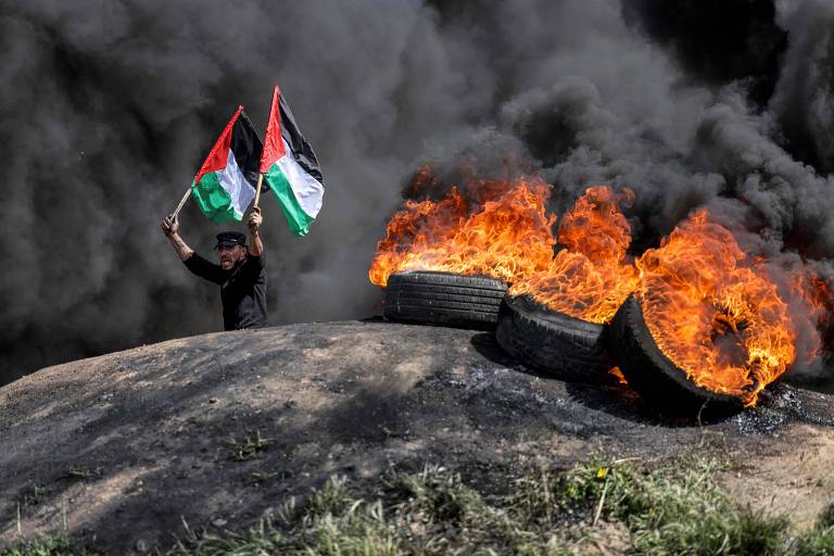Homem ergue bandeiras da Palestina em meio a pneus em chamas em protesto contra invasão da mesquita de Al-Aqsa na fronteira de Israel com a Faixa de Gaza
