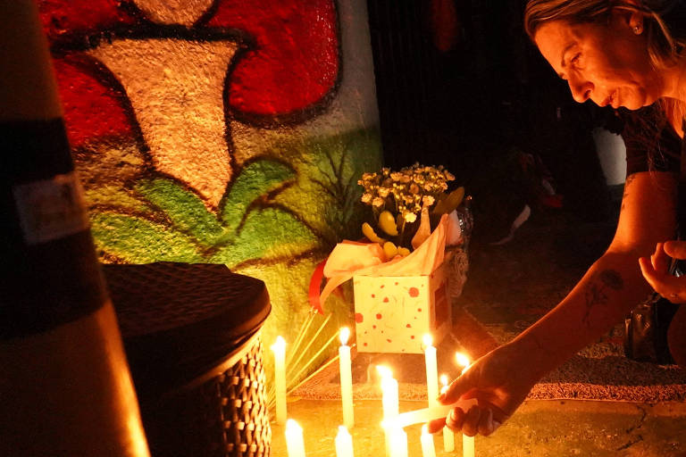 Mulher ajoelhada ao lado de um menino acende uma vela no chão, entre várias outras velas já acesas; a foto foi feita à noite 