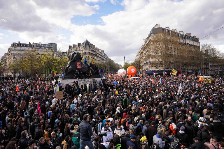 Multidão de pessoas reunidas na Place Denfert-Rochereau durante protesto; na imagem, estão construções ao fundo e diversas pessoas reunidas no promeiro plano, algumas levam balões vermelhos e bandeiras  