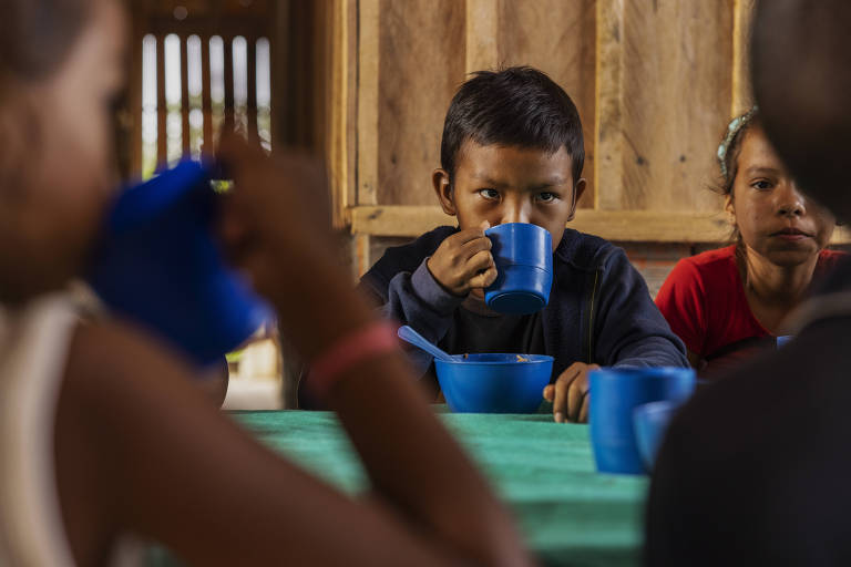 Menino sentado a uma mesa em sala com paredes de tábua toma bebida de uma caneca azul; ao seu lado estão outras crianças