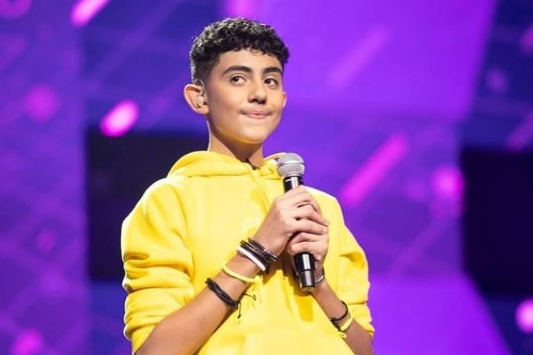 Em foto colorida, adolescente com casaco amarelo sorri enquanto segura o microfone com as duas mãos em uma apresentação