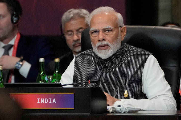 Homem indiano grisalho de barba, camisa branca e colete cinza atrá de placa que diz Índia
