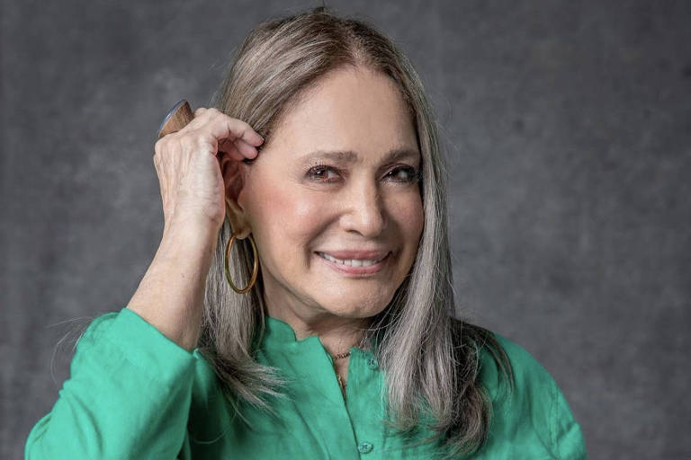 A atriz Susana Vieira, 80, aparece de cabelos grisalhos, vestindo camisa verde, sorrindo e com a mão segurando o cabelo atrás da orelha