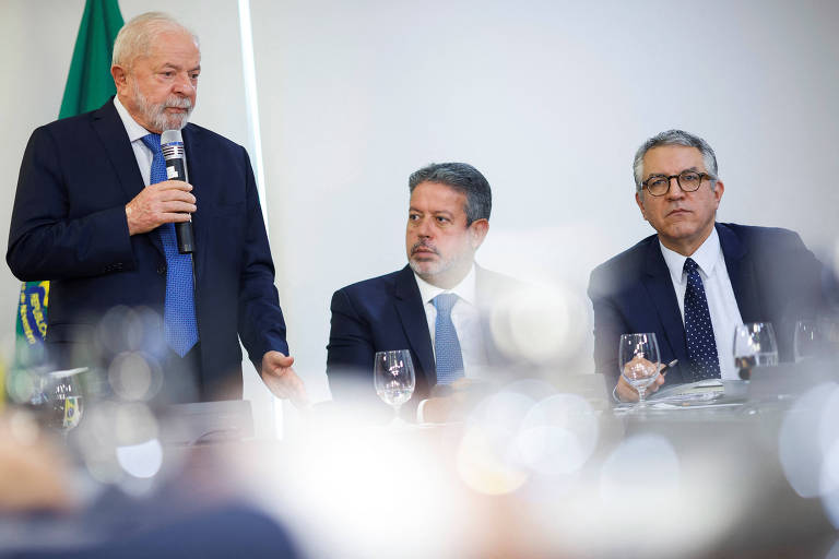 Homem de cabelo e barba grisalhos, de terno escuro e gravata azul e com microfone na mão, fala de pé ao lado de dois homens sentados, de terno e gravata escuros; ao fundo está uma bandeira do Brasil, e a imagem tem reflexos de luz